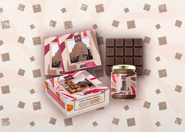 Présentation des produits en chocolat artisanal lyonnais de la collection Héritage : deux tablettes de chocolats carrées dans leur emballage, une tablette sans emballage, un coffret de tablettes de chocolat carré au caramel et un pot en verre de pâte à tartiner, l'emballage de chaque produit présente des motifs pop et colorés bordeaux et marron et une illustration d'un célèbre bâtiment lyonnais dans le style BD. L'arrière-plan de la photo est beige clair effet vieilli à motifs de petites tablettes de chocolat de plusieurs tailles.